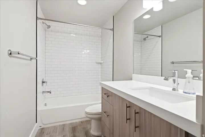 Bathroom featuring a white subway tile tub surround, chrome fixtures, a wood-finish vanity with a white countertop, and wood-finish flooring