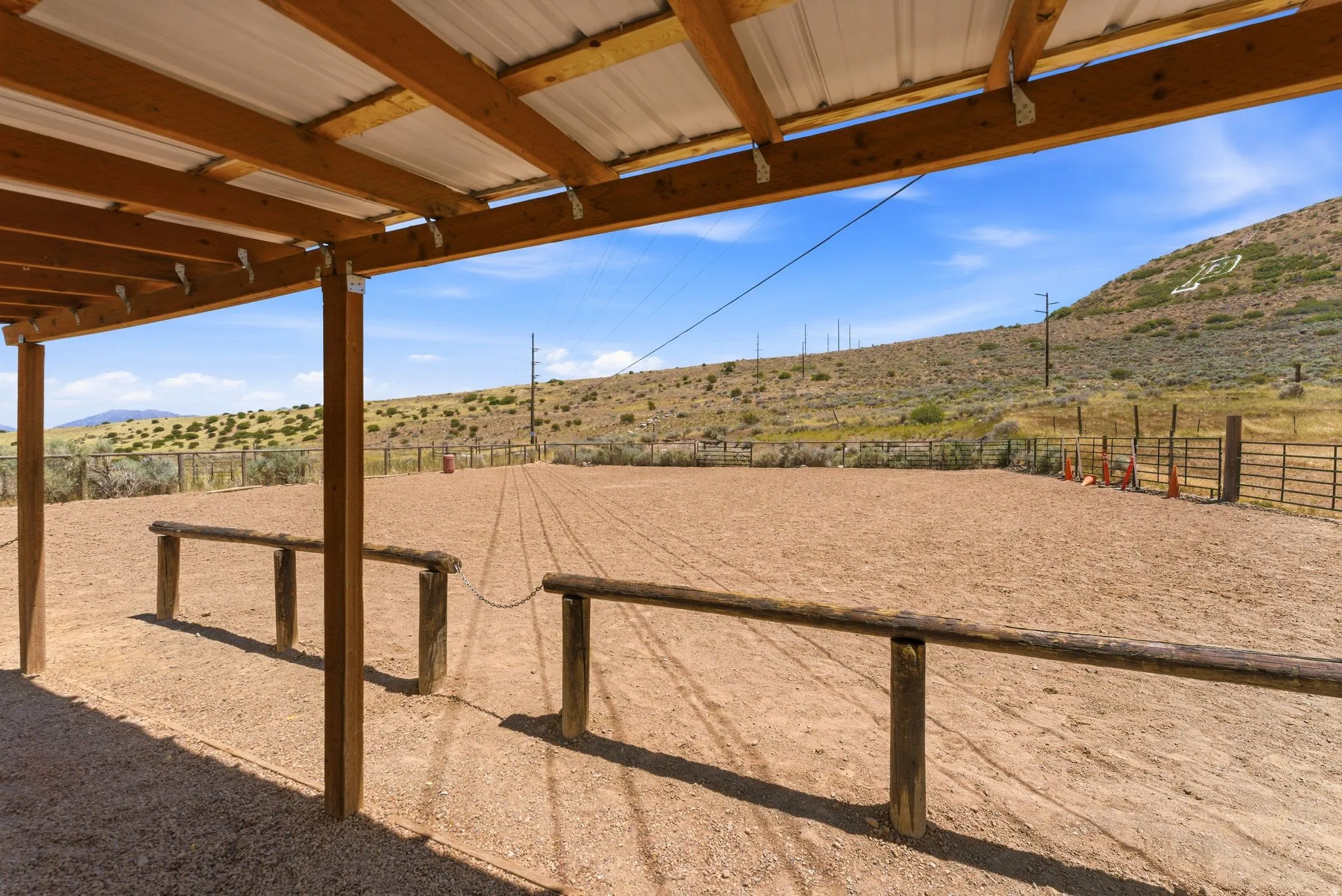 Covered outdoor structure with wood beams and a metal roof, overlooking a spacious riding arena with a gravel surface