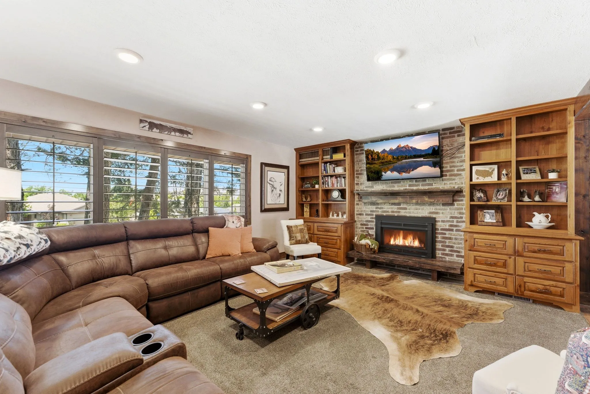 Living room featuring a brick fireplace with a wood mantle, built-in wooden cabinetry, recessed lighting, and large windows with plantation shutters