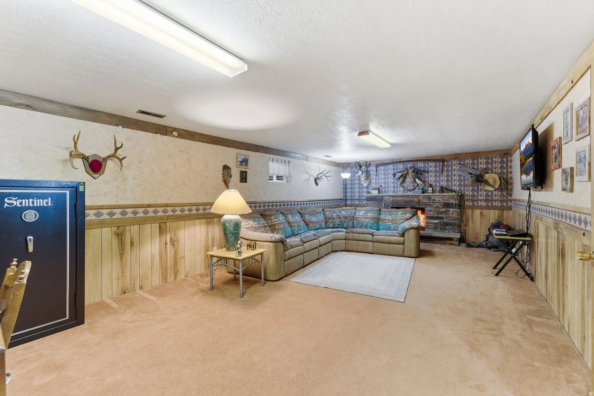 Spacious lower-level room featuring an integrated stone fireplace, wood-finish wall paneling, and neutral carpeting