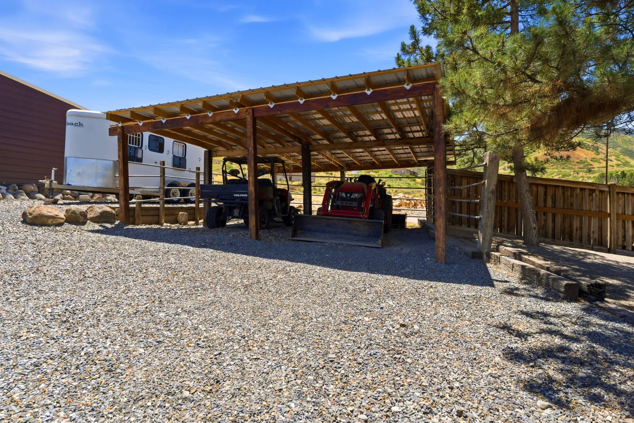 Wood-frame covered structure with a corrugated metal roof, gravel flooring, and a natural wood fence