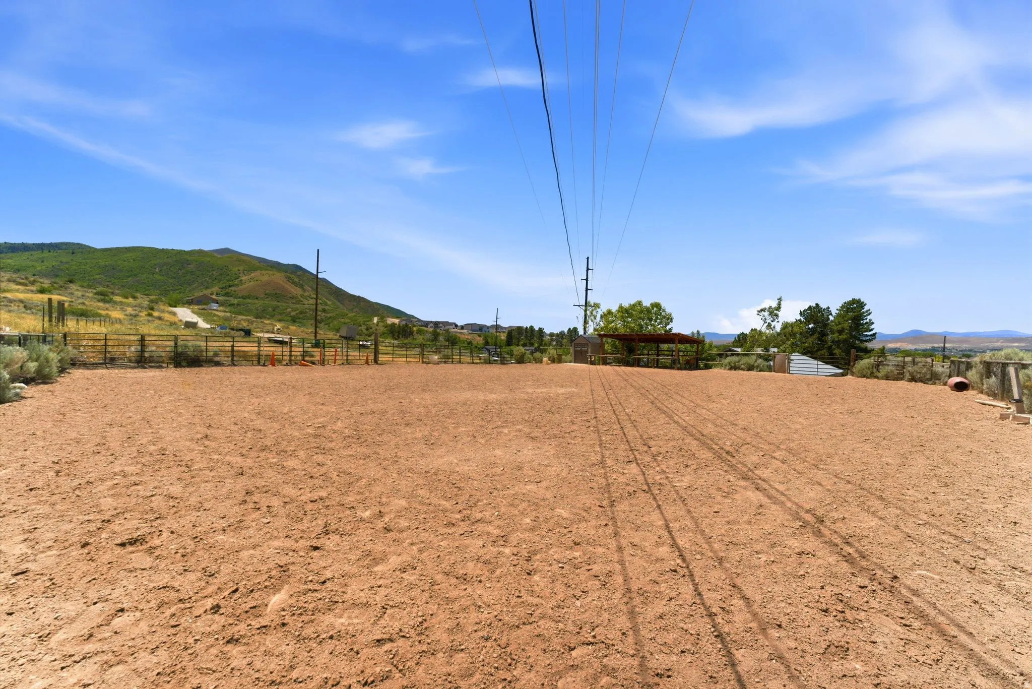 Open-air riding arena with perimeter fencing, a covered structure, and distant mountain backdrops