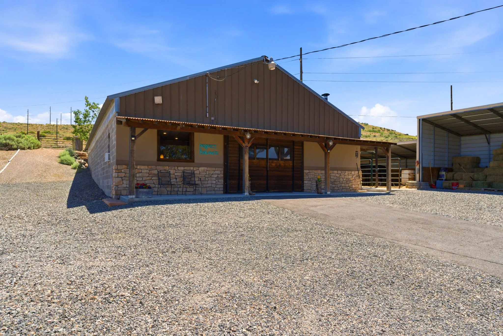 Single-story building with a brown metal roof and siding, stone veneer accents, a covered front porch with wood posts, wood-finish double doors, and a gravel driveway