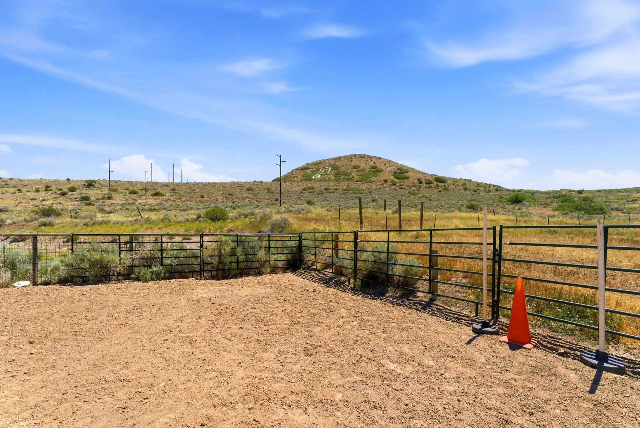 Fenced outdoor area with a dirt surface and metal panel fencing, surrounded by natural grasslands and distant hills