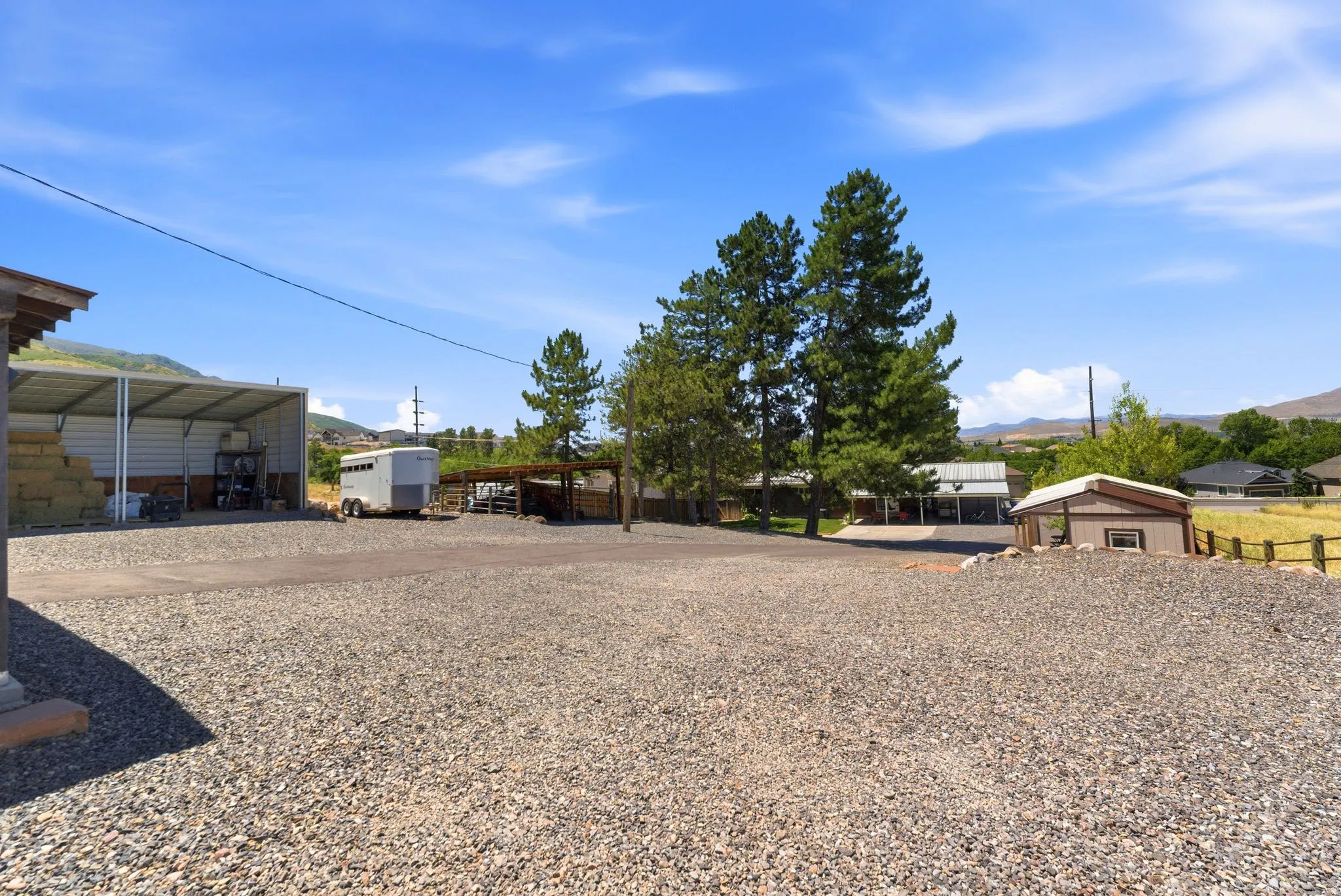 Expansive gravel lot featuring a large open-sided metal structure, mature pine trees, a small shed, and distant mountain scenery