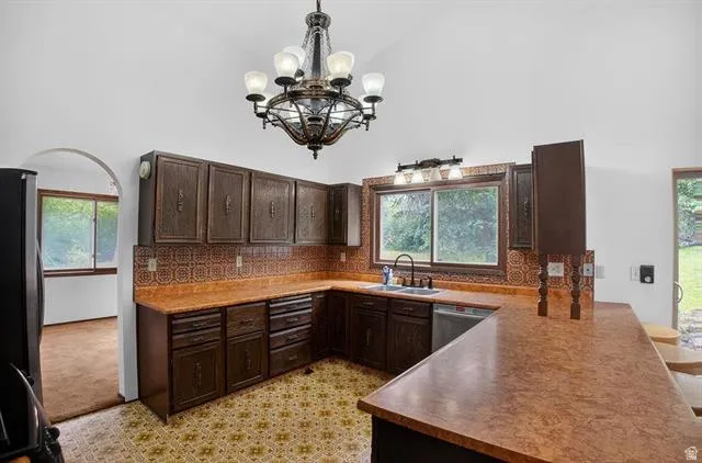 Kitchen featuring vaulted ceilings, a central chandelier, wood-finish cabinetry, a large window overlooking natural scenery, and a built-in dishwasher