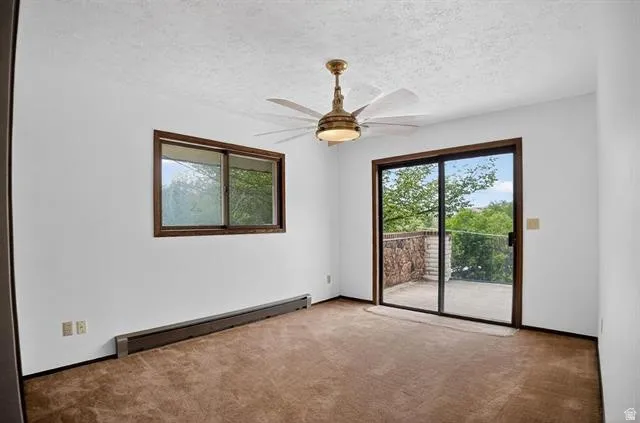 Carpeted room featuring a ceiling fan with light, a sliding glass door providing access to an outdoor patio with a stone wall, and a horizontal window with dark wood trim