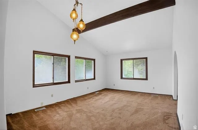 Spacious room featuring vaulted ceilings with an exposed beam, three large windows with dark trim, a decorative three-light pendant fixture, and an arched entryway