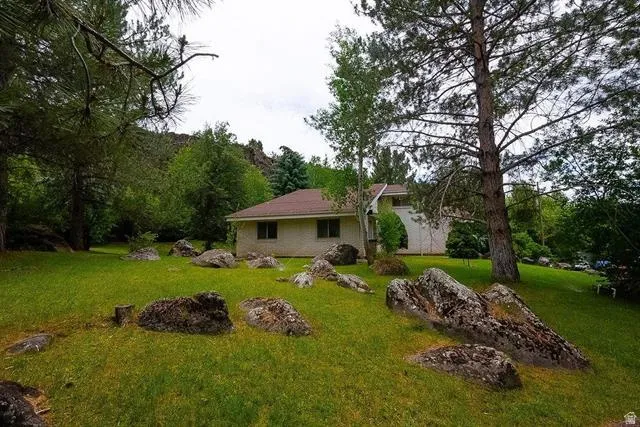 Expansive green lawn with scattered natural rock formations, mature pine trees, and a distant tan single-story residence with a brown roof