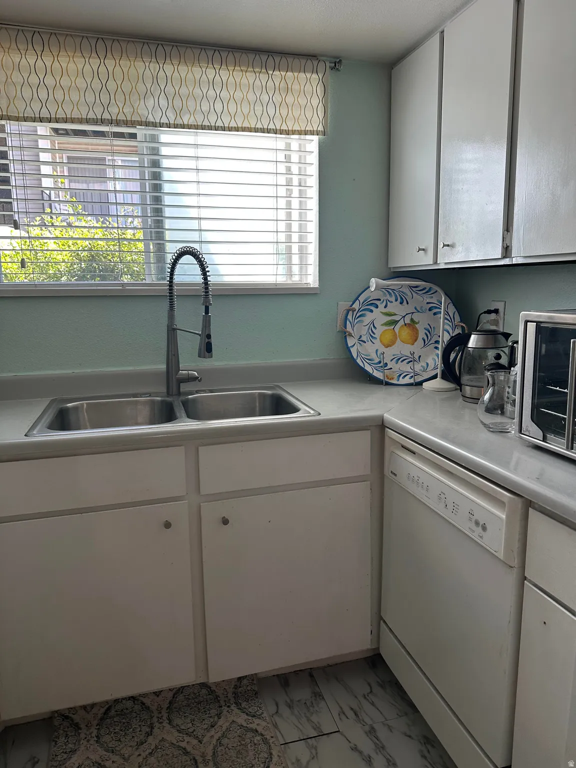 Kitchen with a double basin stainless steel sink, gooseneck faucet, light countertops, white cabinetry, and a white dishwasher