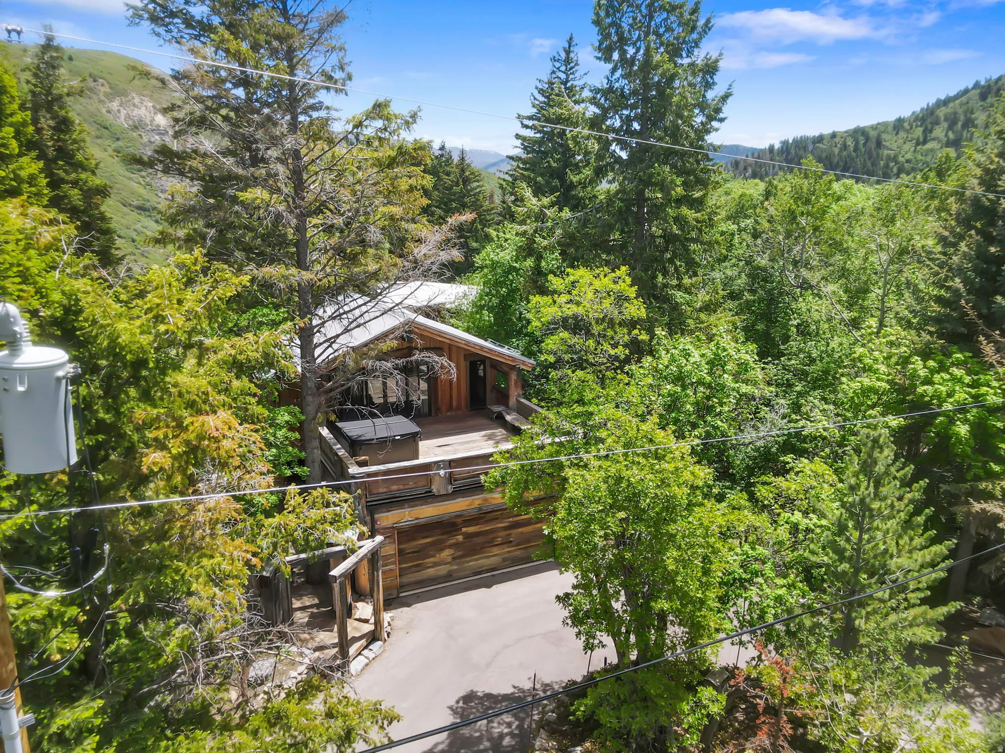 Wood-sided cabin exterior with a large wooden deck and an integrated hot tub, surrounded by mature trees and a paved driveway