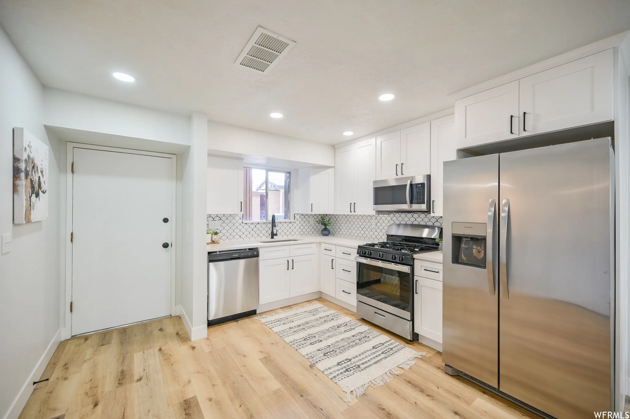 Modern kitchen featuring white shaker cabinetry with black hardware, stainless steel appliances, a geometric tile backsplash, and light wood-finish flooring