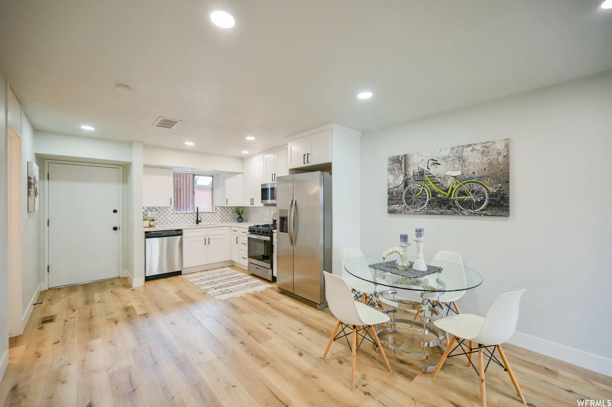 Open-concept kitchen and dining area featuring light wood-finish flooring, white cabinetry, stainless steel appliances, a geometric tile backsplash, and recessed lighting