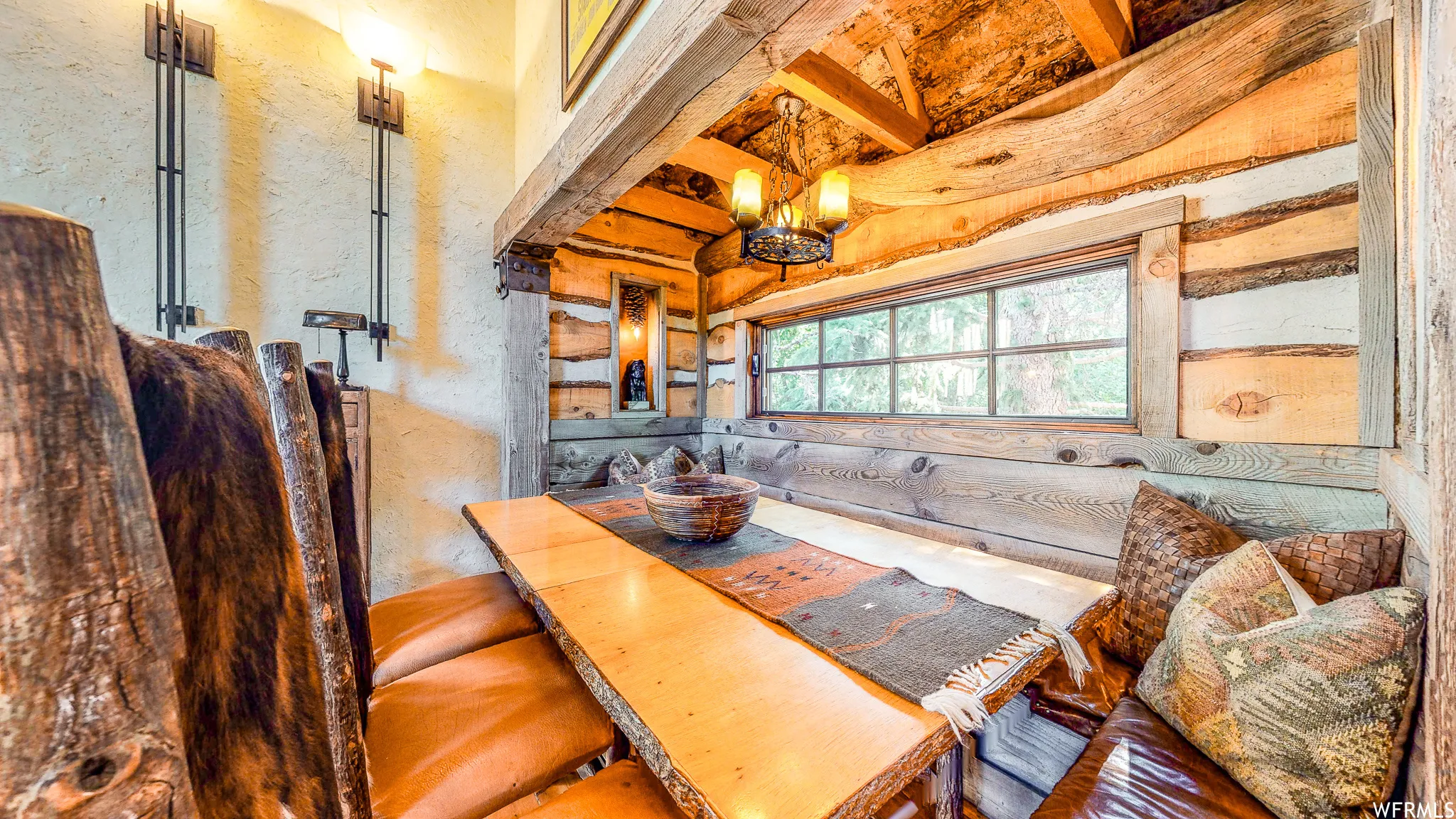 Dining area featuring log construction, a built-in bench with leather cushions, a raw-edge wood table, a window with exterior views, and an ornate metal chandelier