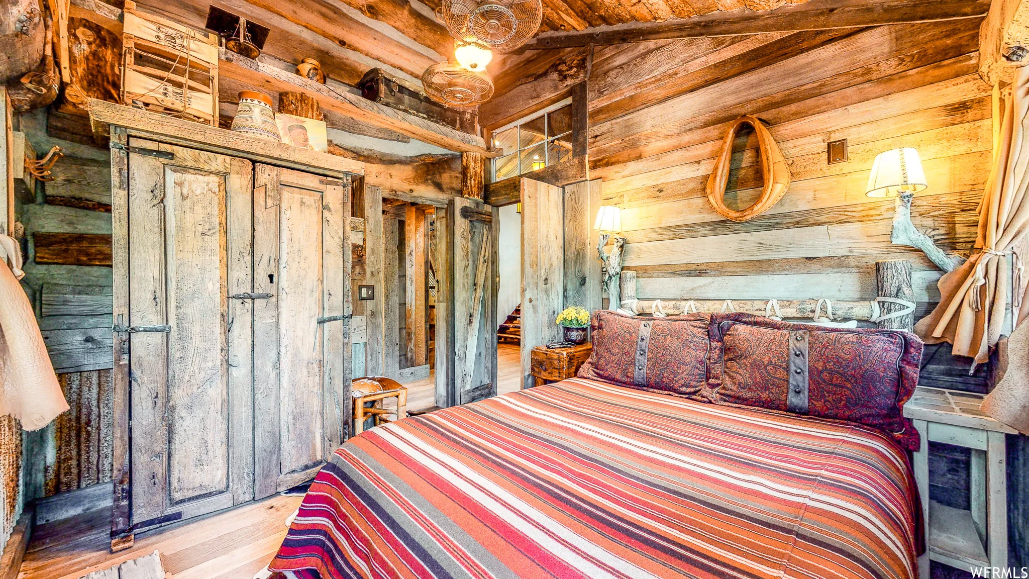 Wood-paneled room featuring a prominent bed with a decorative headboard, two wall-mounted lamps with fabric shades, a rustic wood-finish wardrobe with double doors, and a wood-finish ceiling with exposed beams