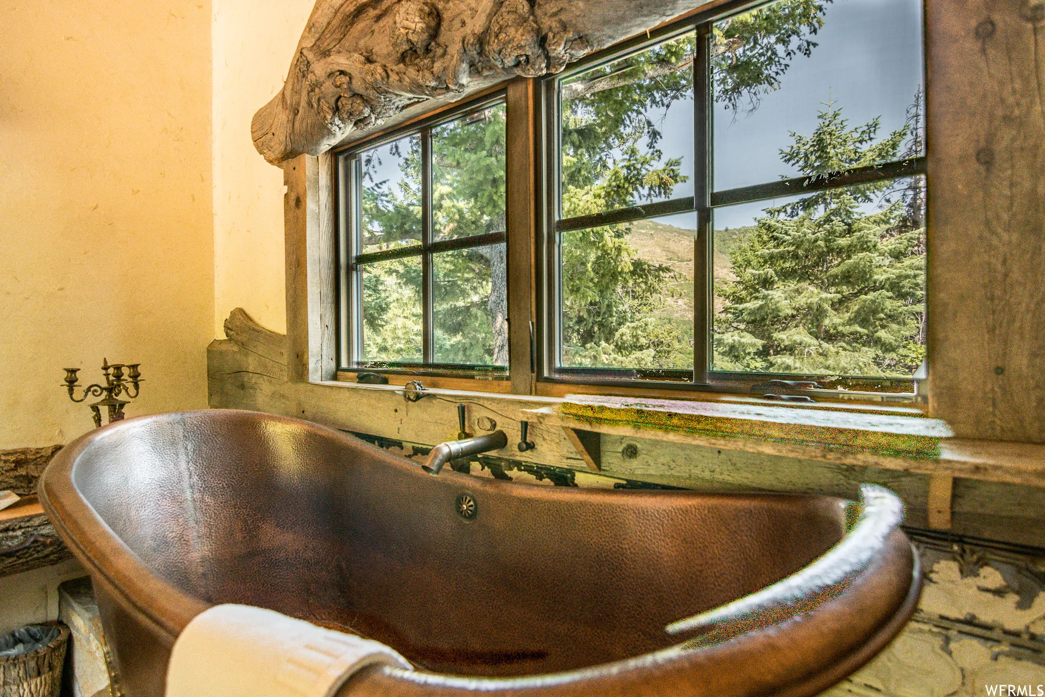 Freestanding copper bathtub with a wall-mounted faucet, set against a rustic window frame with exposed wood elements, textured wall finish, and a natural outdoor backdrop