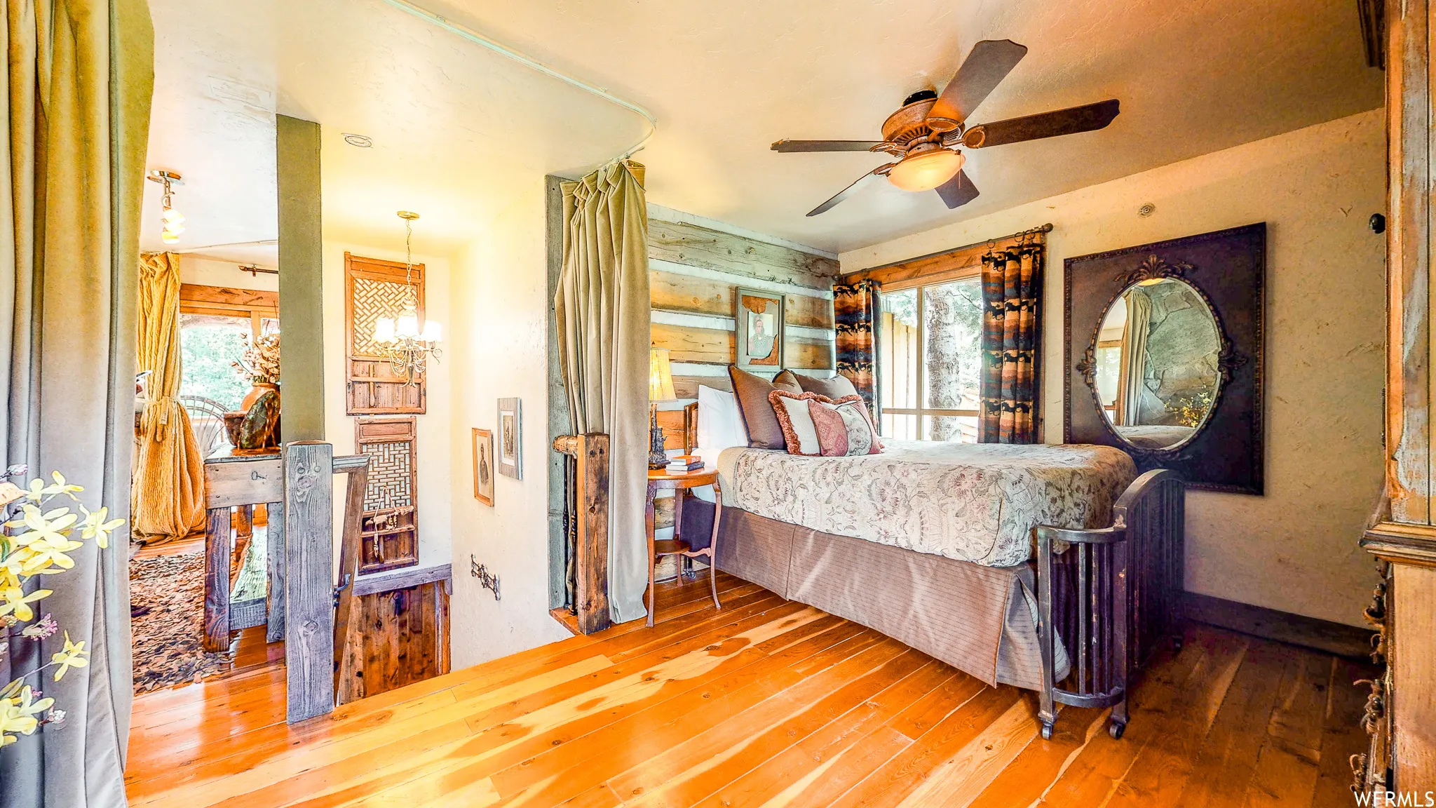 Loft bedroom featuring wood-finish flooring, a ceiling fan with integrated lighting, wood paneled accent wall, and double windows with patterned drapes