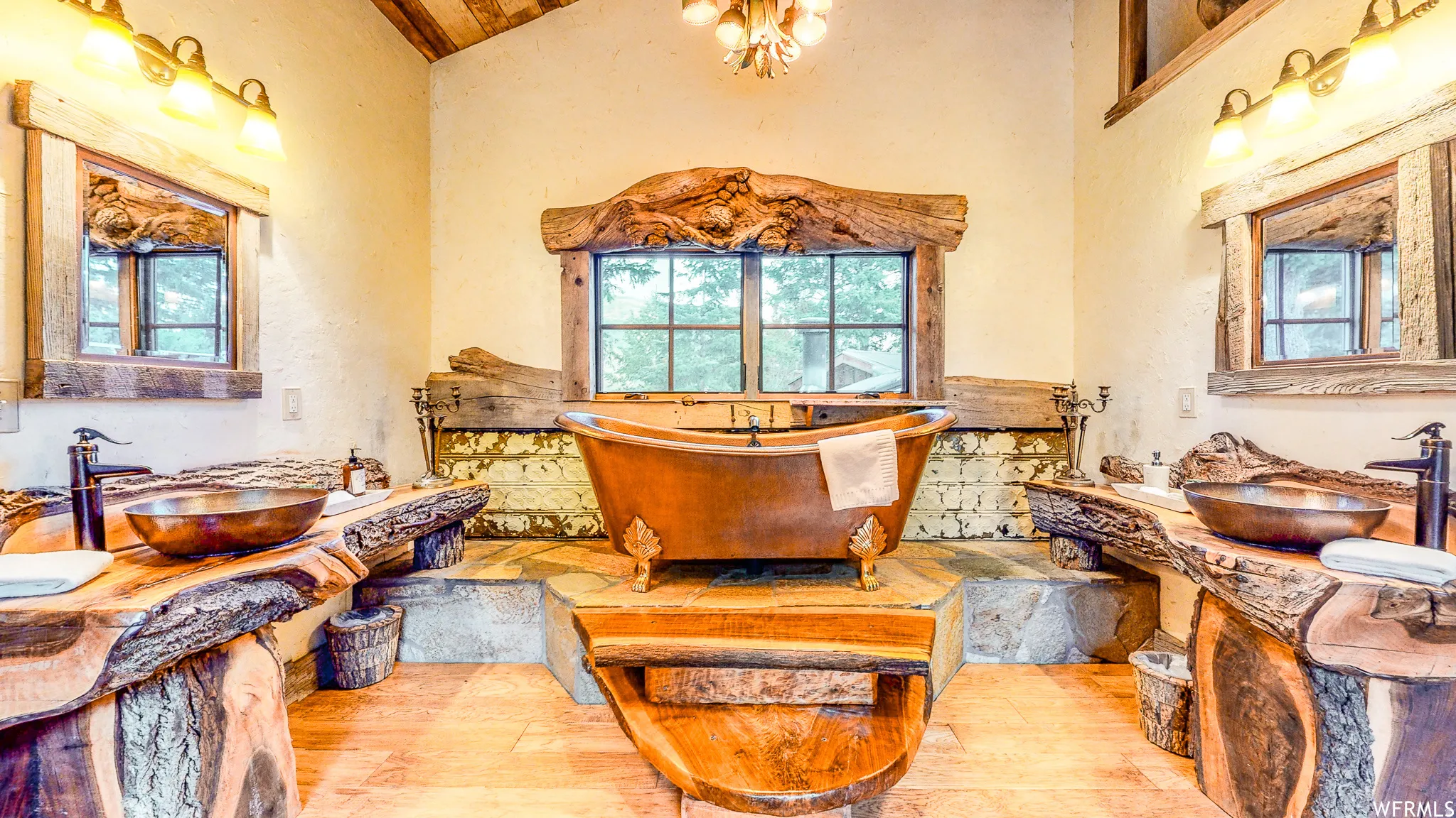 Distinguished bathroom featuring a copper-tone freestanding bathtub, two distinct wood-slab vanities with integrated vessel sinks, two framed mirrors, and rustic wood window trim