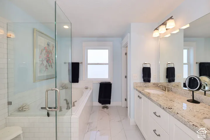 Bathroom featuring a frameless glass shower enclosure, a built-in jetted tub, a dual vanity with a light-colored speckled countertop, white cabinetry, and a large mirror with integrated lighting