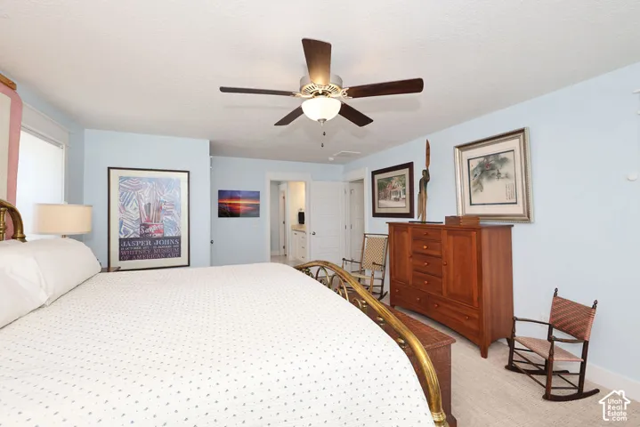 Spacious room featuring a ceiling fan with light fixture, light-colored carpeting, multiple windows, and a doorway leading to an additional space with a vanity