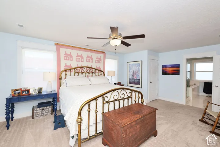 Carpeted bedroom featuring a ceiling fan with integrated lighting, two windows with white trim, and a doorway opening to a bathroom with tile flooring and a window