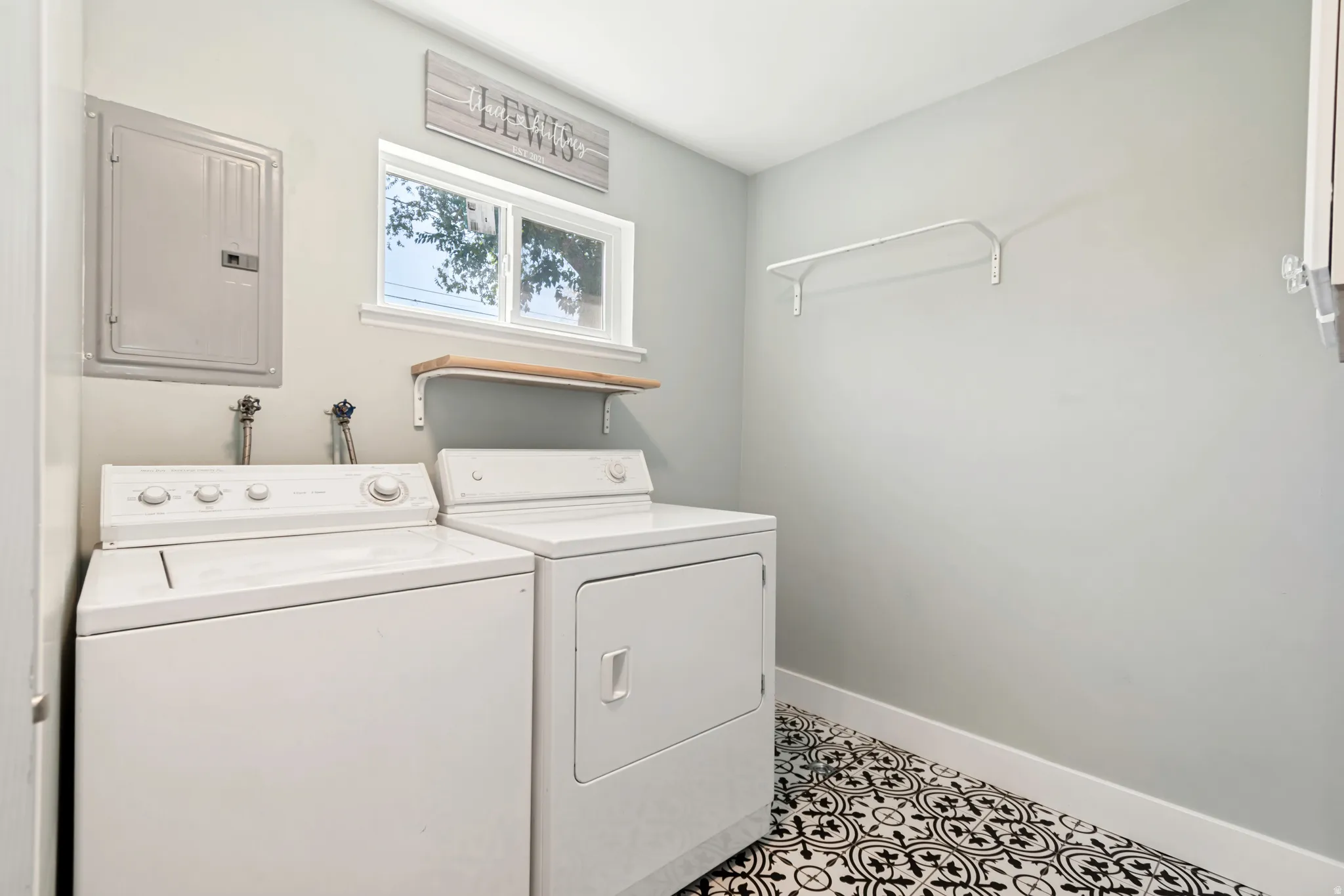 Laundry area featuring patterned floor tiles, light-toned walls, a window, and a wall-mounted shelf