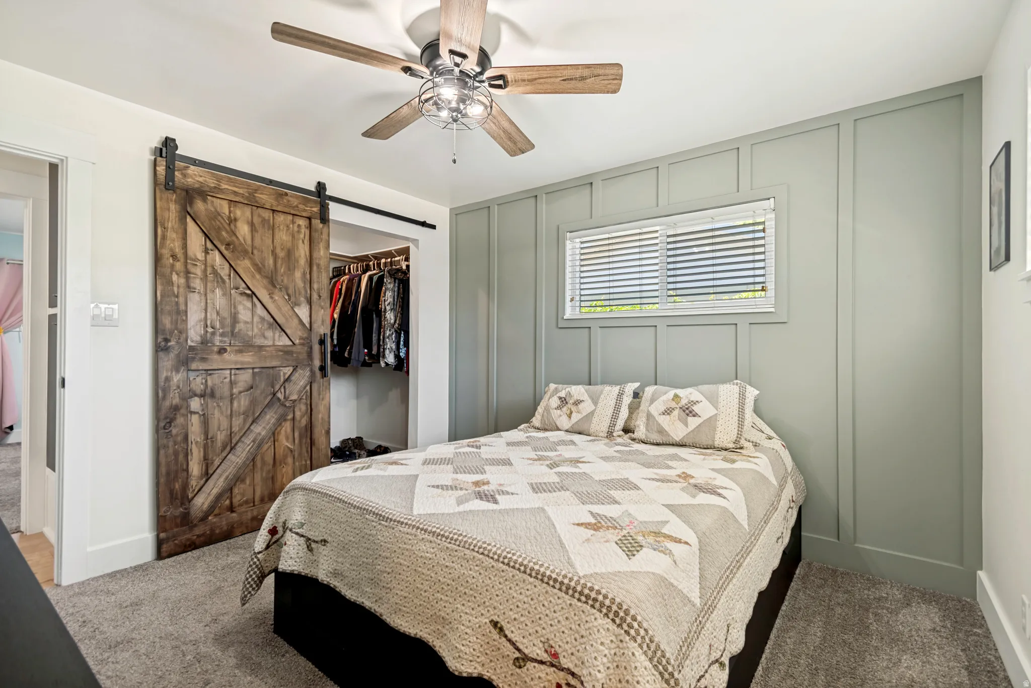 Bedroom featuring wainscoting accent wall, window with blinds, and ceiling fan and custom barn door