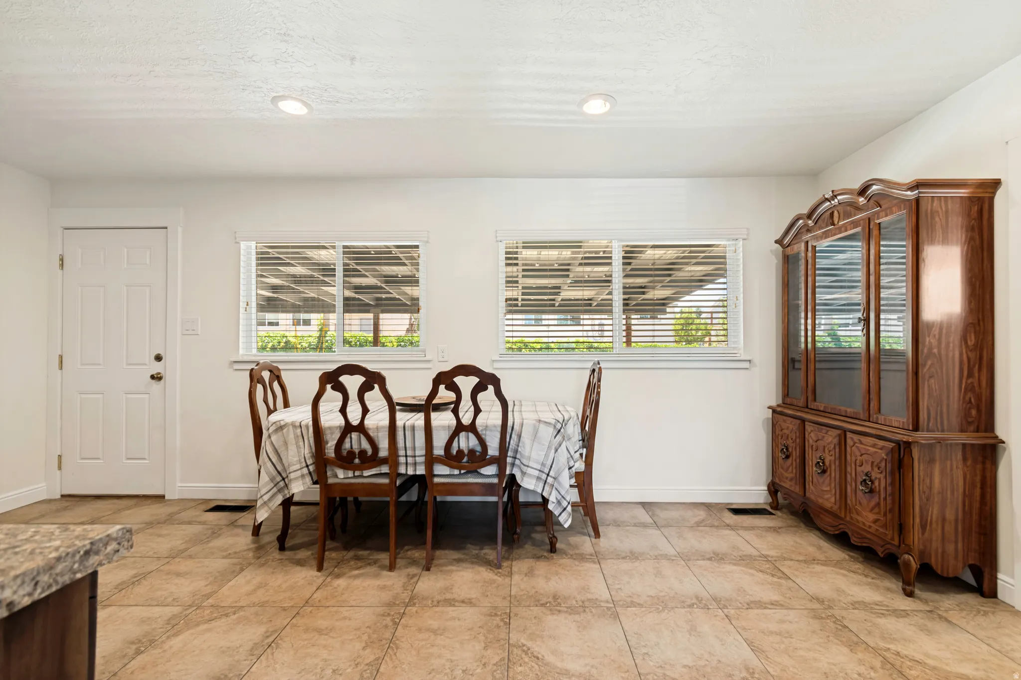Tiled flooring extending into a dining area featuring two windows with horizontal blinds, recessed lighting, and a white paneled door