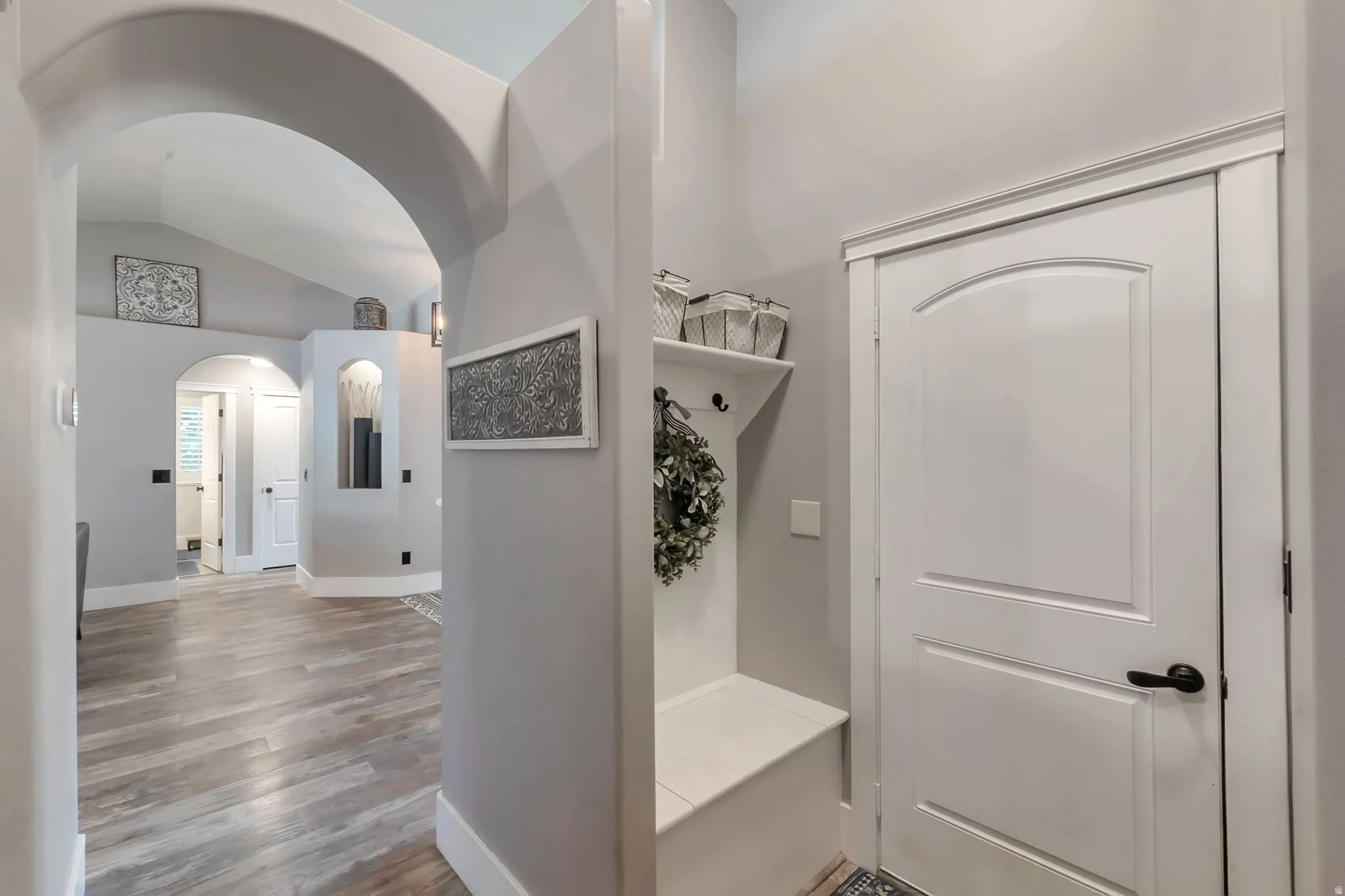Arched entryway leading to an interior hallway with wood-finish flooring, a built-in entryway bench with shelving, and white interior doors with dark hardware