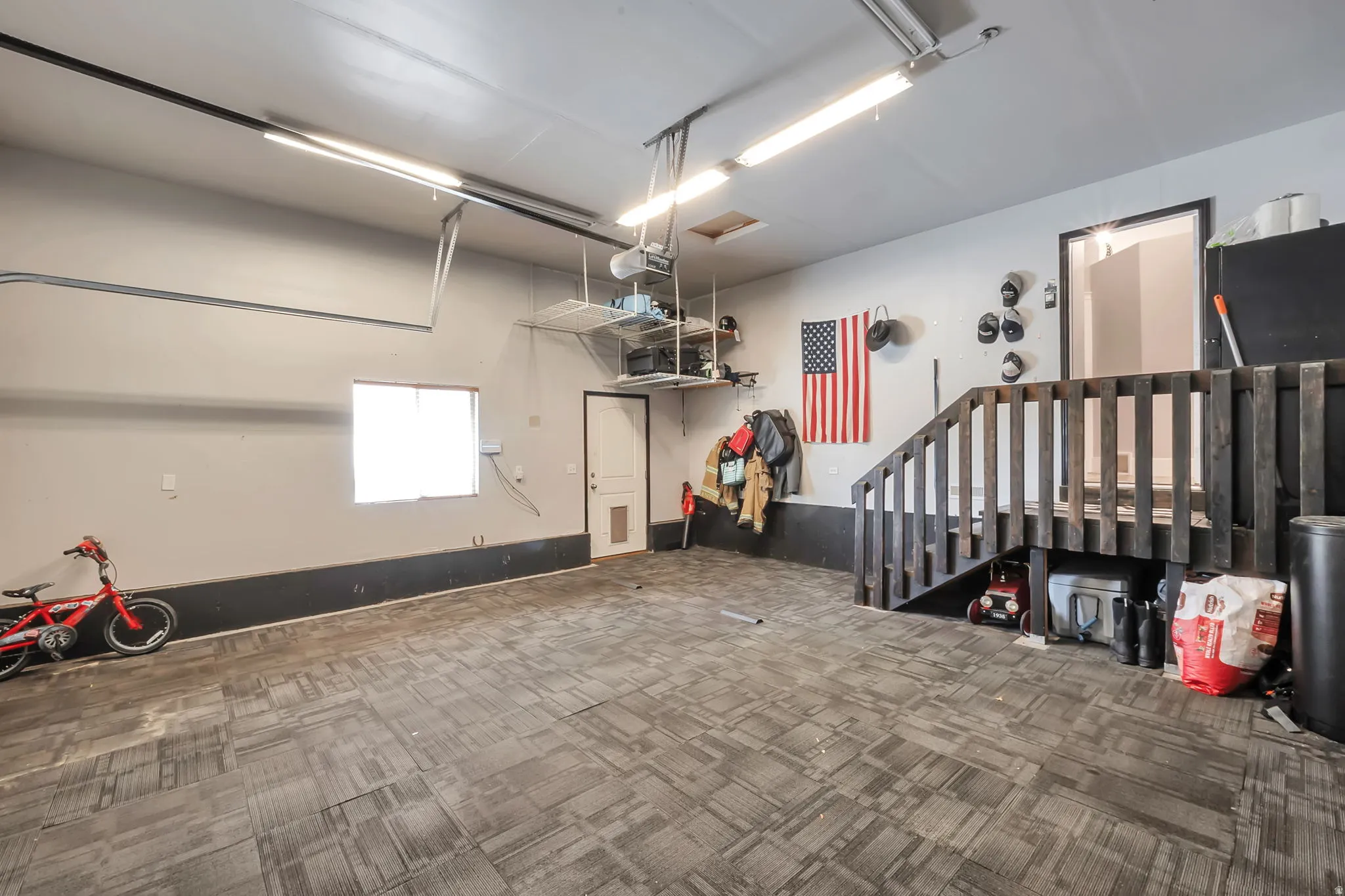 Spacious garage featuring patterned carpet tile flooring, a window providing natural light, and a built-in wooden staircase with a railing