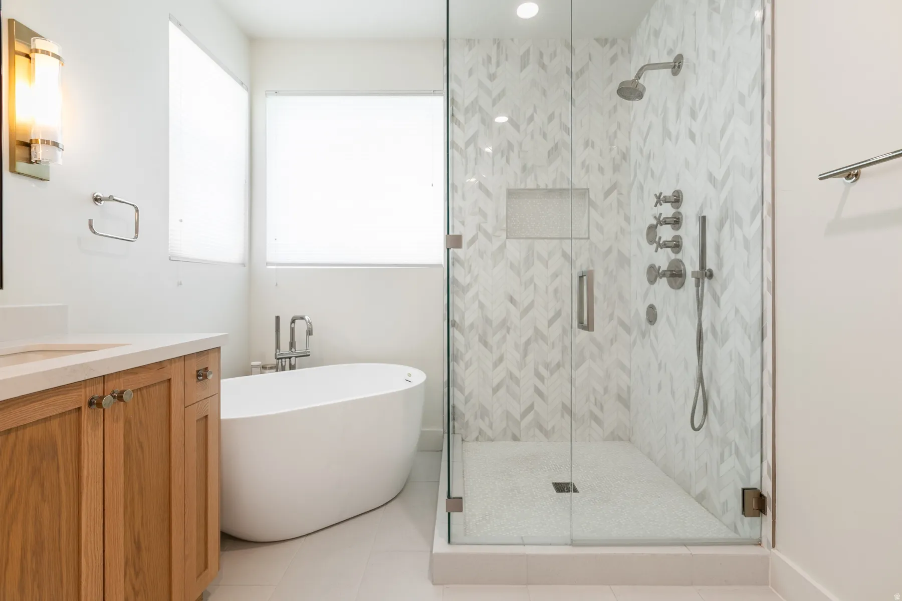 Contemporary bathroom featuring a freestanding soaking tub, a frameless glass shower enclosure with herringbone tile, a recessed shower niche, and a vanity with natural wood-finish cabinetry and a light-toned countertop