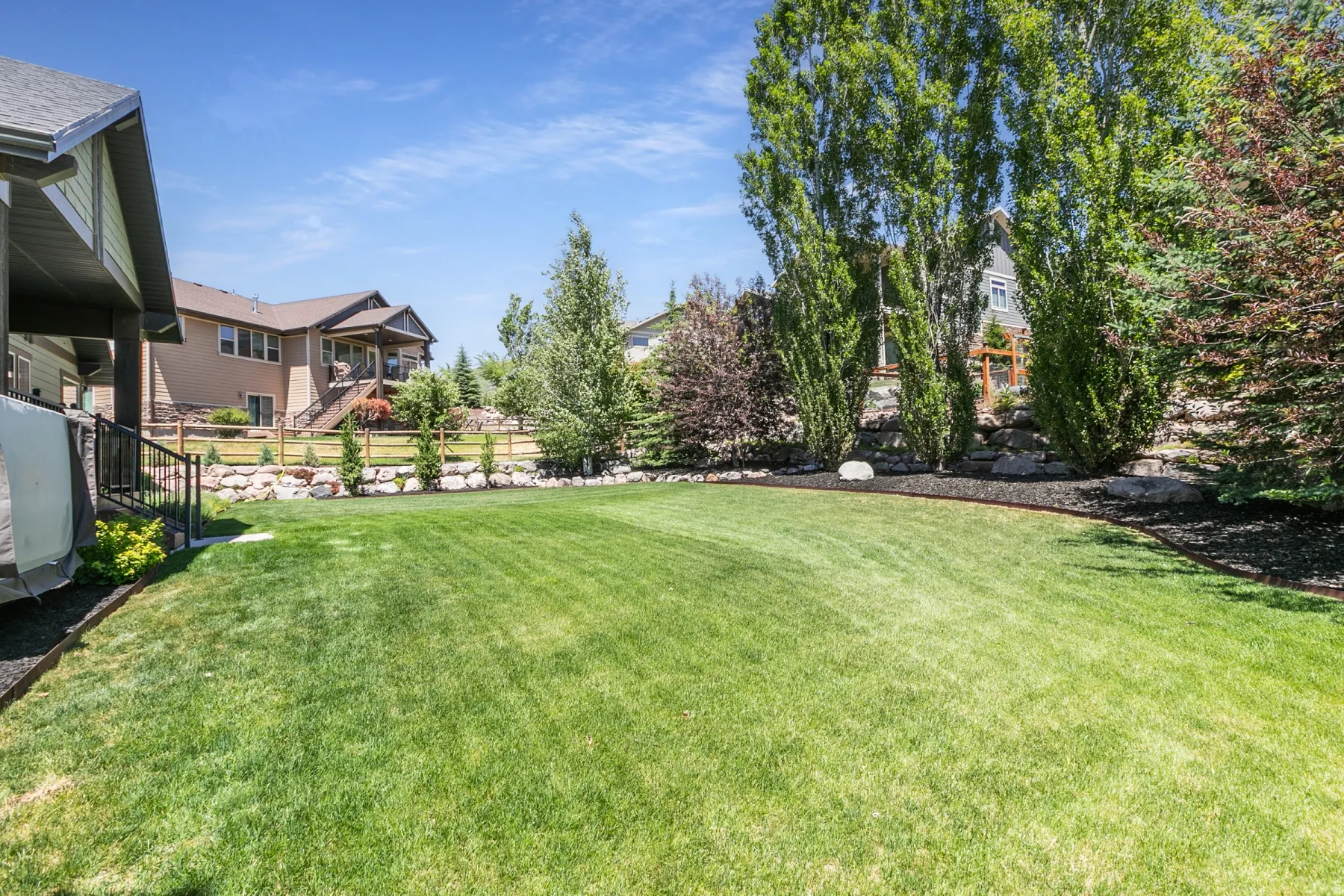 Expansive green lawn with mature trees and shrubbery, featuring a decorative rock retaining wall, mulched landscape beds, and partial views of neighboring properties