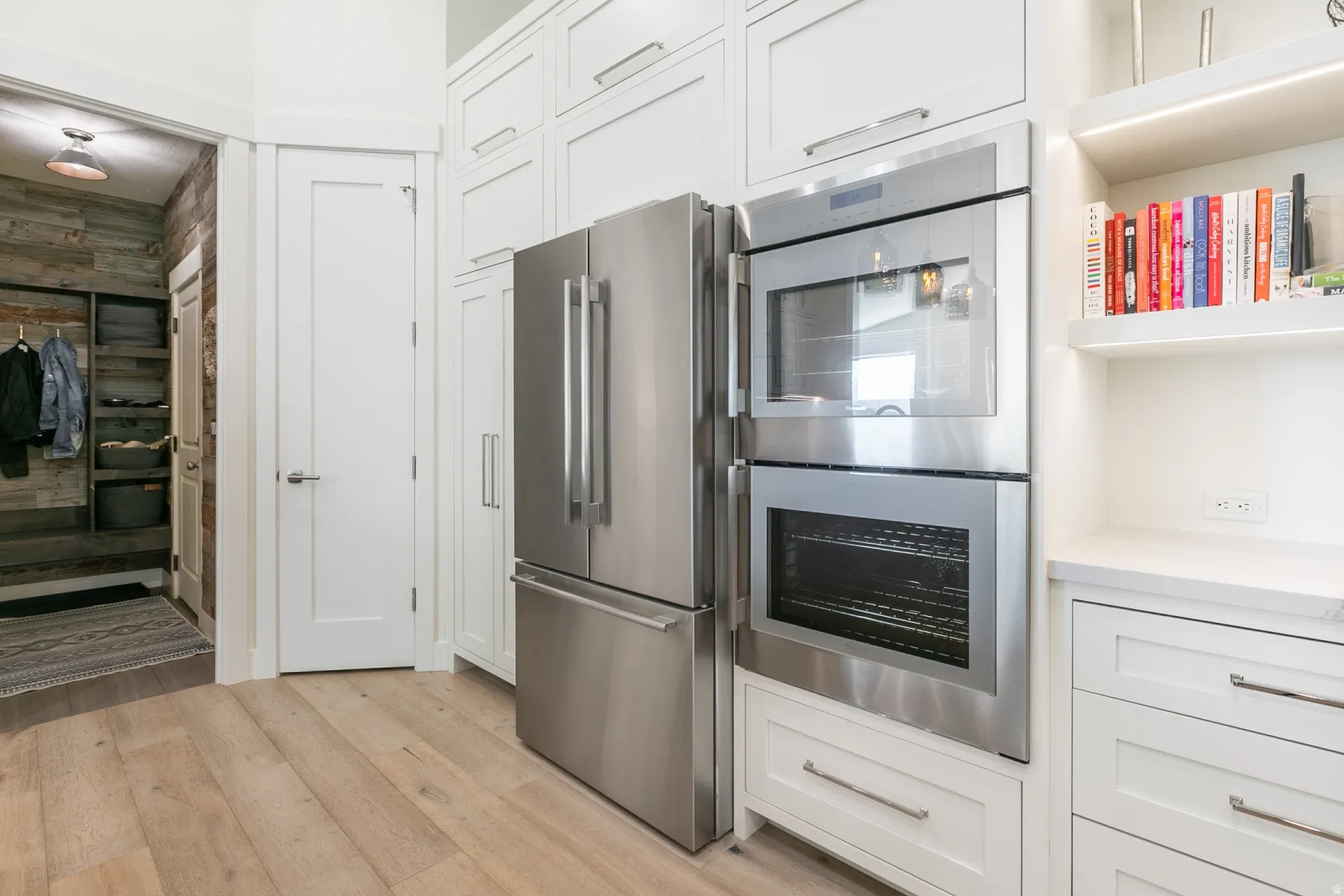 Stainless steel refrigerator and double wall ovens integrated into white cabinetry with matching hardware, wood-finish flooring, and built-in shelving with concealed lighting