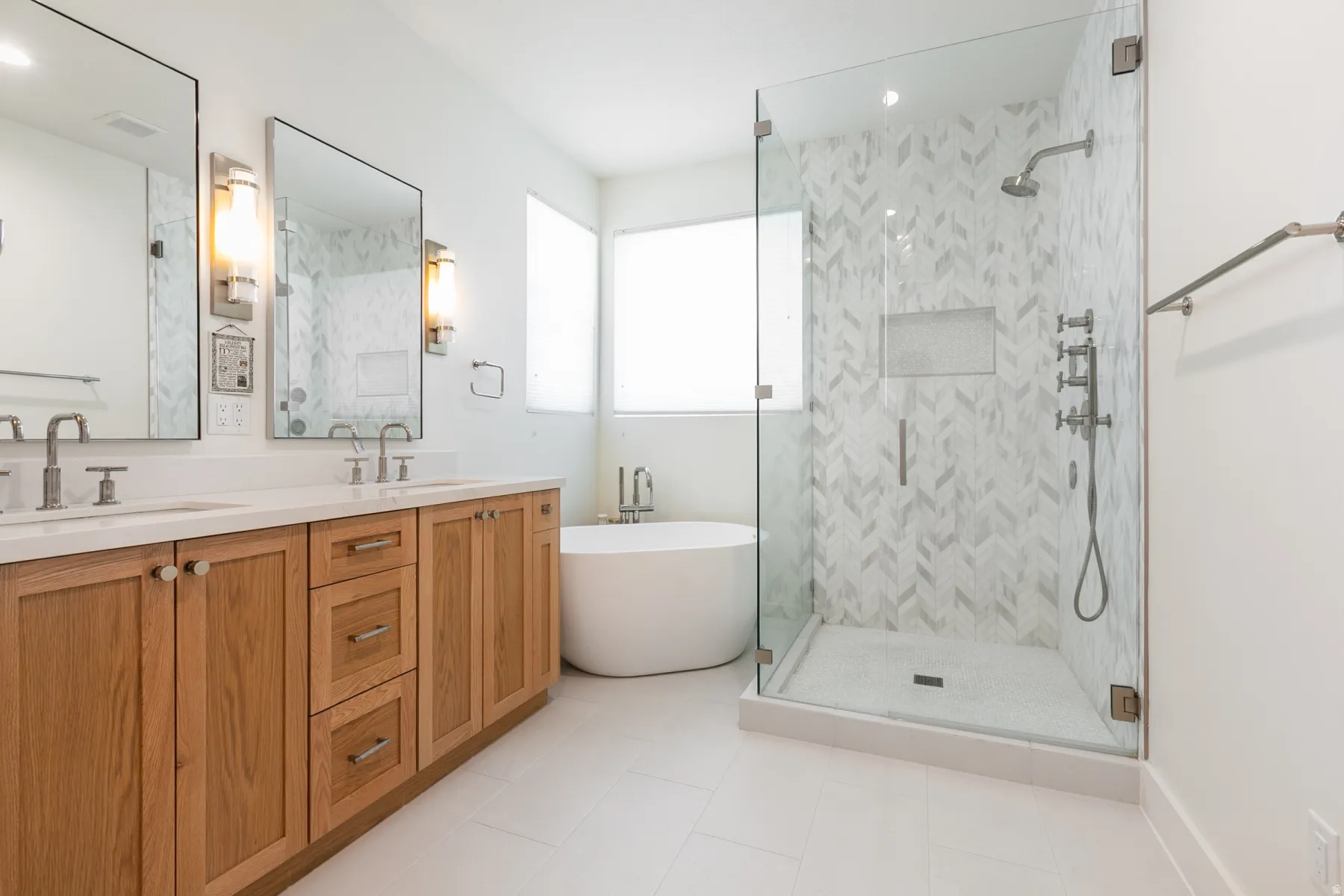 Modern bathroom featuring a freestanding tub, glass-enclosed shower with chevron tile, dual vanity with wood cabinetry, and rectangular mirrors with sconce lighting