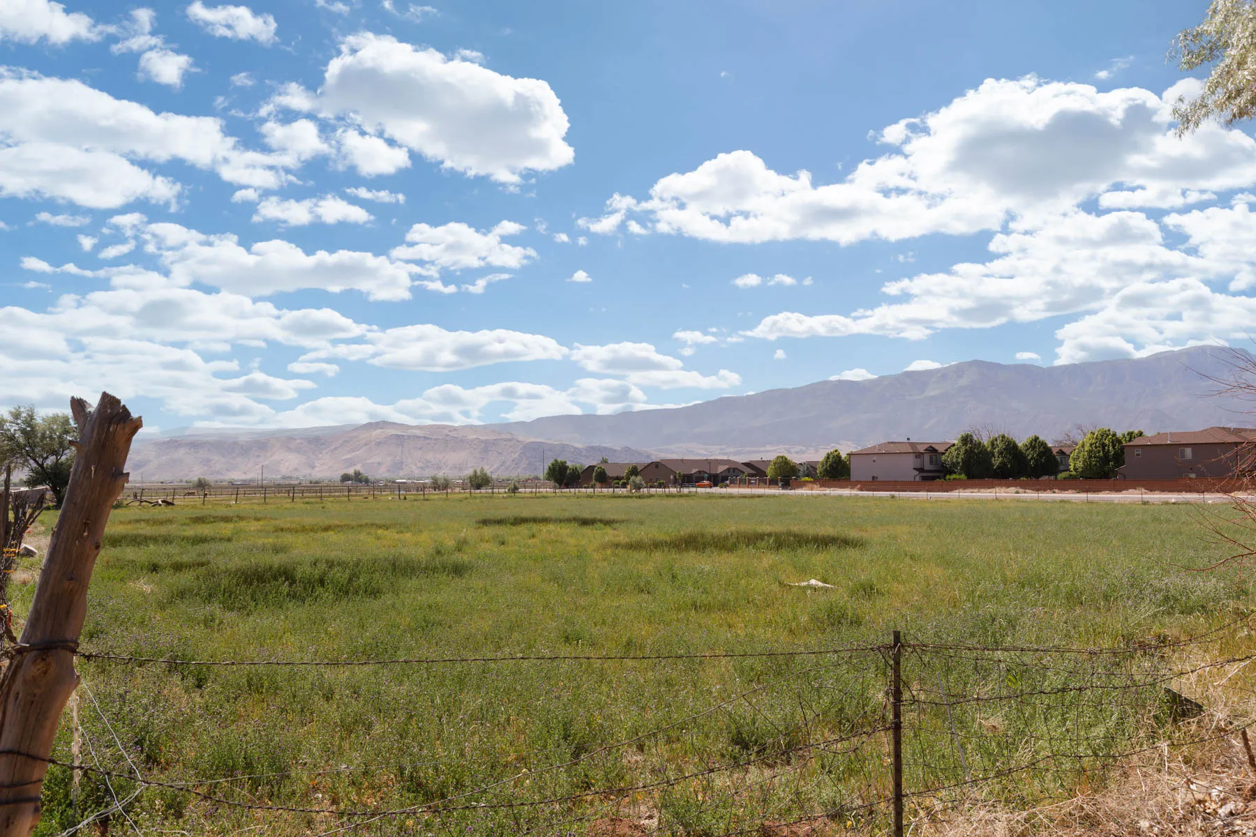Expansive green space featuring a wire fence, distant residential structures, and a prominent mountain range backdrop