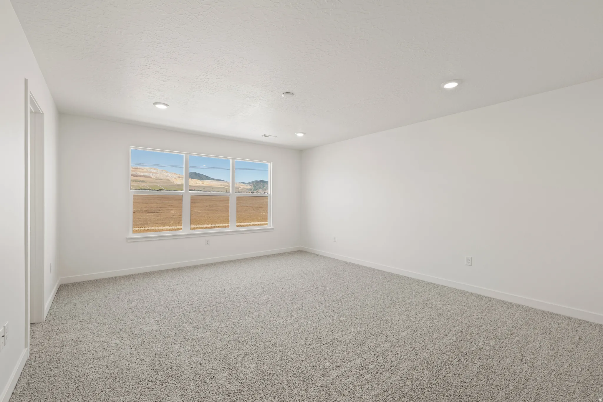 Spacious room featuring light gray carpeting, crisp white walls, recessed ceiling lighting, and a multi-pane window showcasing a scenic natural landscape