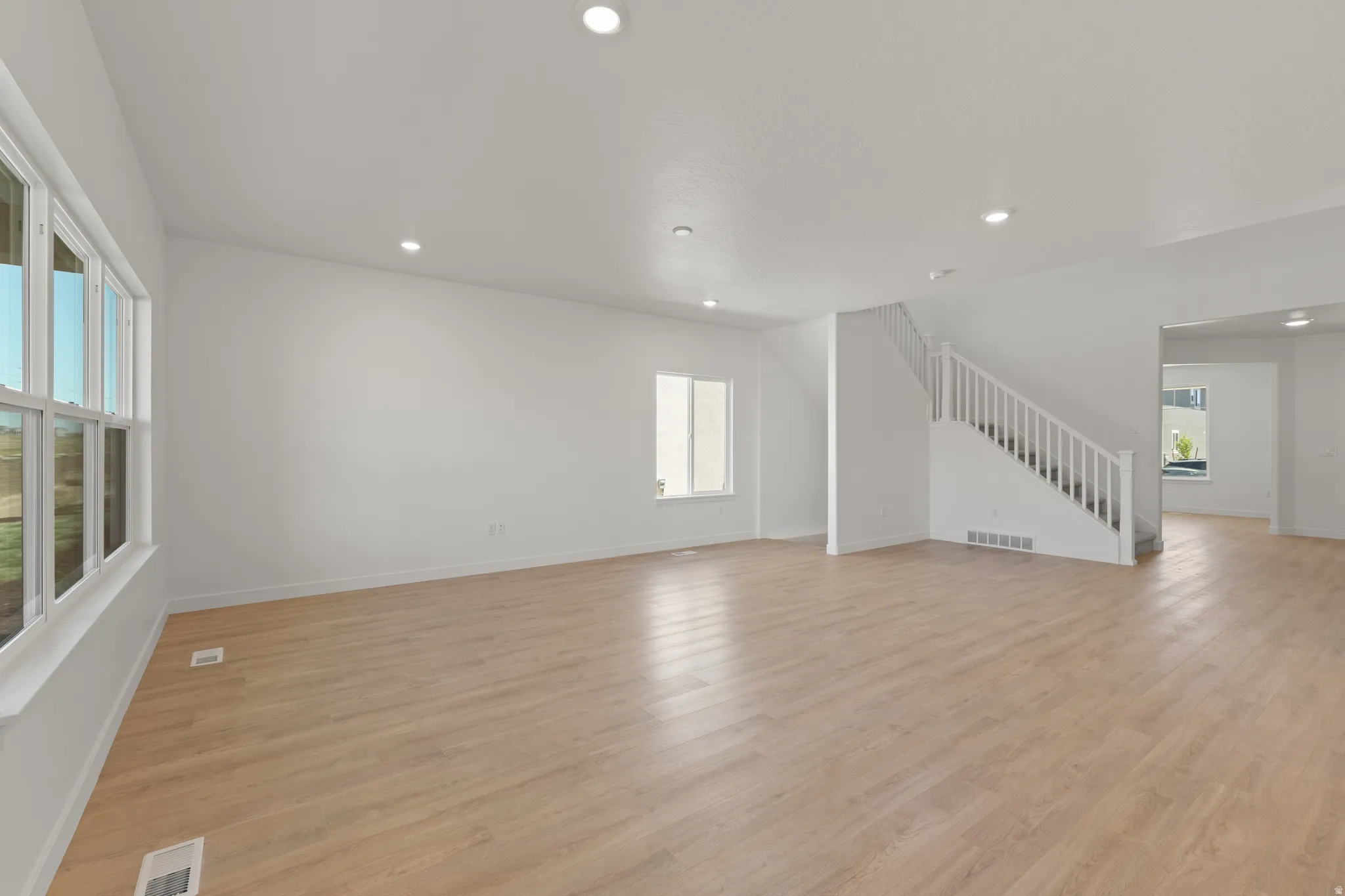 Spacious room featuring wood-finish flooring, ample natural light from multiple windows, recessed lighting, and a white staircase with balusters