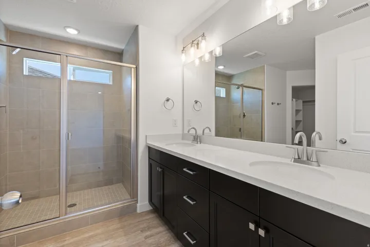 Bathroom featuring a spacious glass-enclosed shower with tile surround, a double vanity with stone-finish countertops, wood-finish flooring, and contemporary lighting fixtures