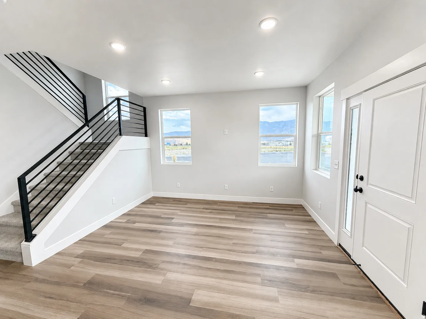 Spacious open area featuring wood-finish flooring, a modern black horizontal bar railing staircase with carpeted treads, recessed ceiling lighting, and multiple windows
