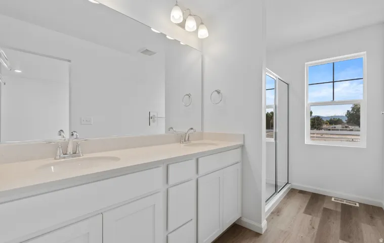 Double vanity with integrated sinks and chrome faucets, light-toned countertop, wood-finish flooring, glass-enclosed shower, and a large window