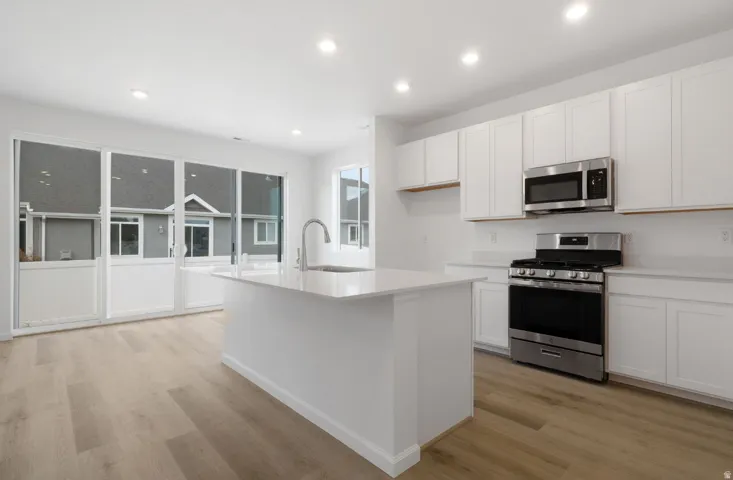 Recessed lighting illuminates the kitchen featuring white cabinetry, a center island with sink, and stainless steel appliances