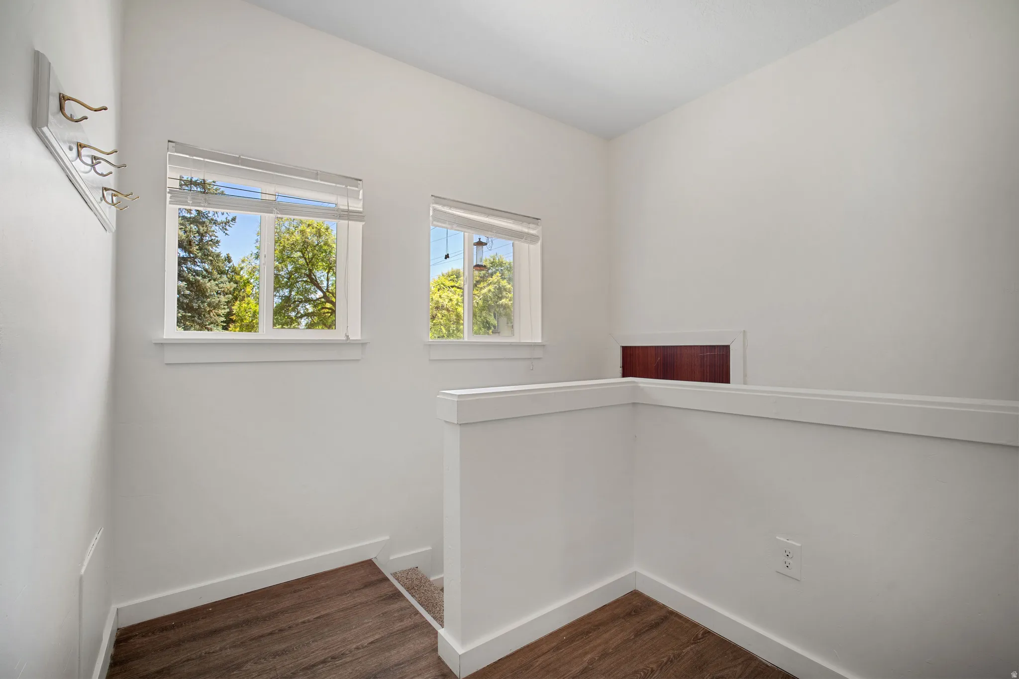 Bright loft space featuring two windows with horizontal blinds, wood-finish flooring, white walls, and a partial low wall