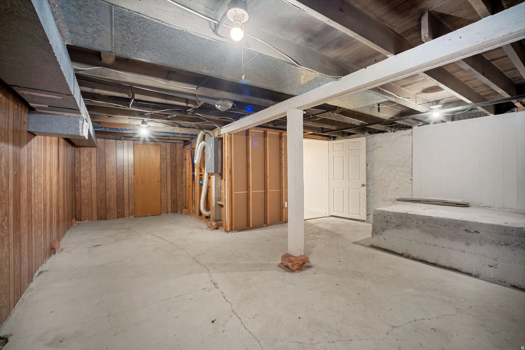 Spacious unfinished lower level featuring exposed ceiling joists, concrete flooring, wood-panel wall accents, and an accessible doorway