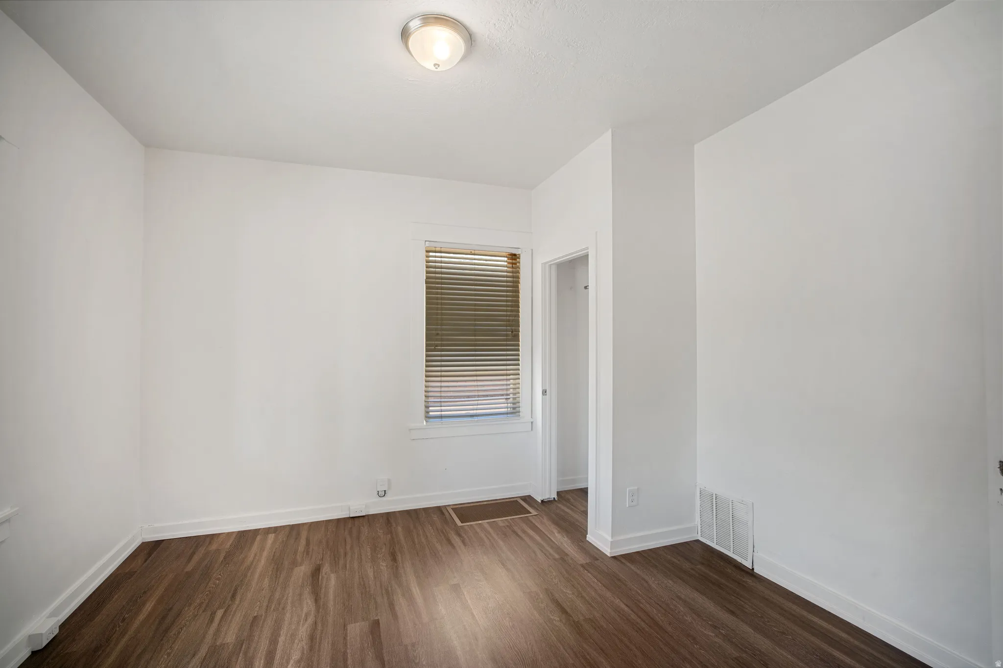 Room featuring white walls, wood-finish flooring, a window with horizontal blinds, a ceiling-mounted light fixture, and an open doorway