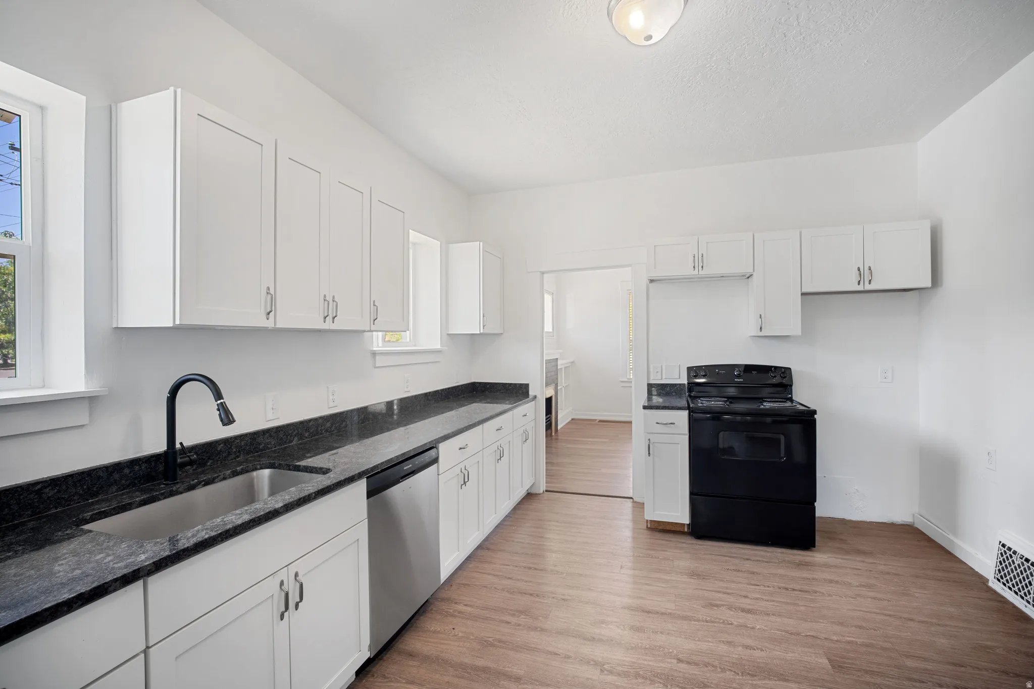 Bright kitchen with white cabinetry, dark stone countertops, a stainless steel sink with a matte black faucet, a stainless steel dishwasher, and a black range