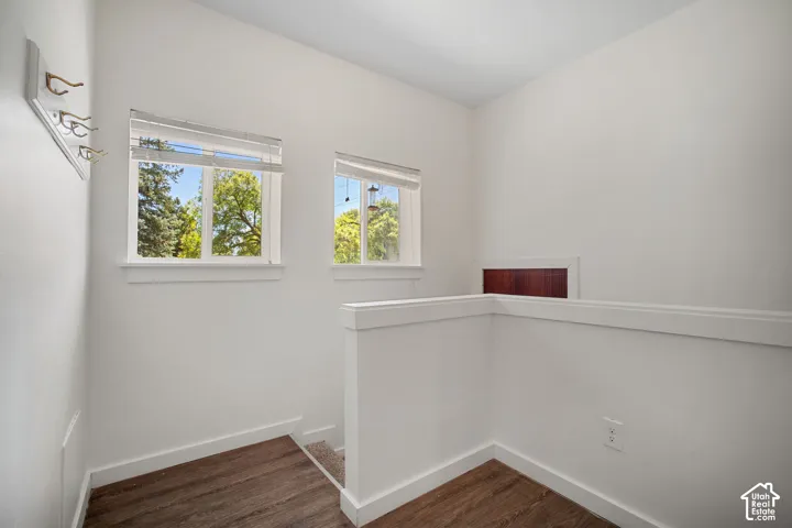 Bright loft space featuring two windows with horizontal blinds, wood-finish flooring, white walls, and a partial low wall