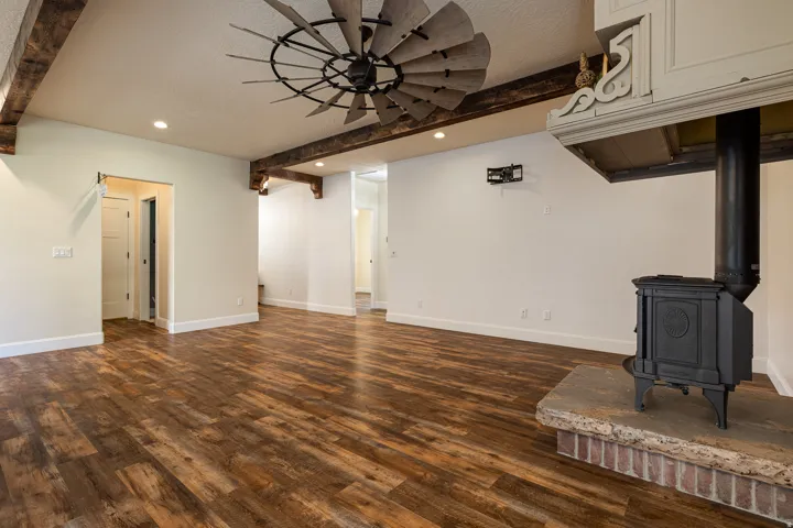 Living area featuring wood-finish flooring, exposed ceiling beams, a decorative ceiling fan, a cast iron wood-burning stove with a stone hearth, and recessed lighting