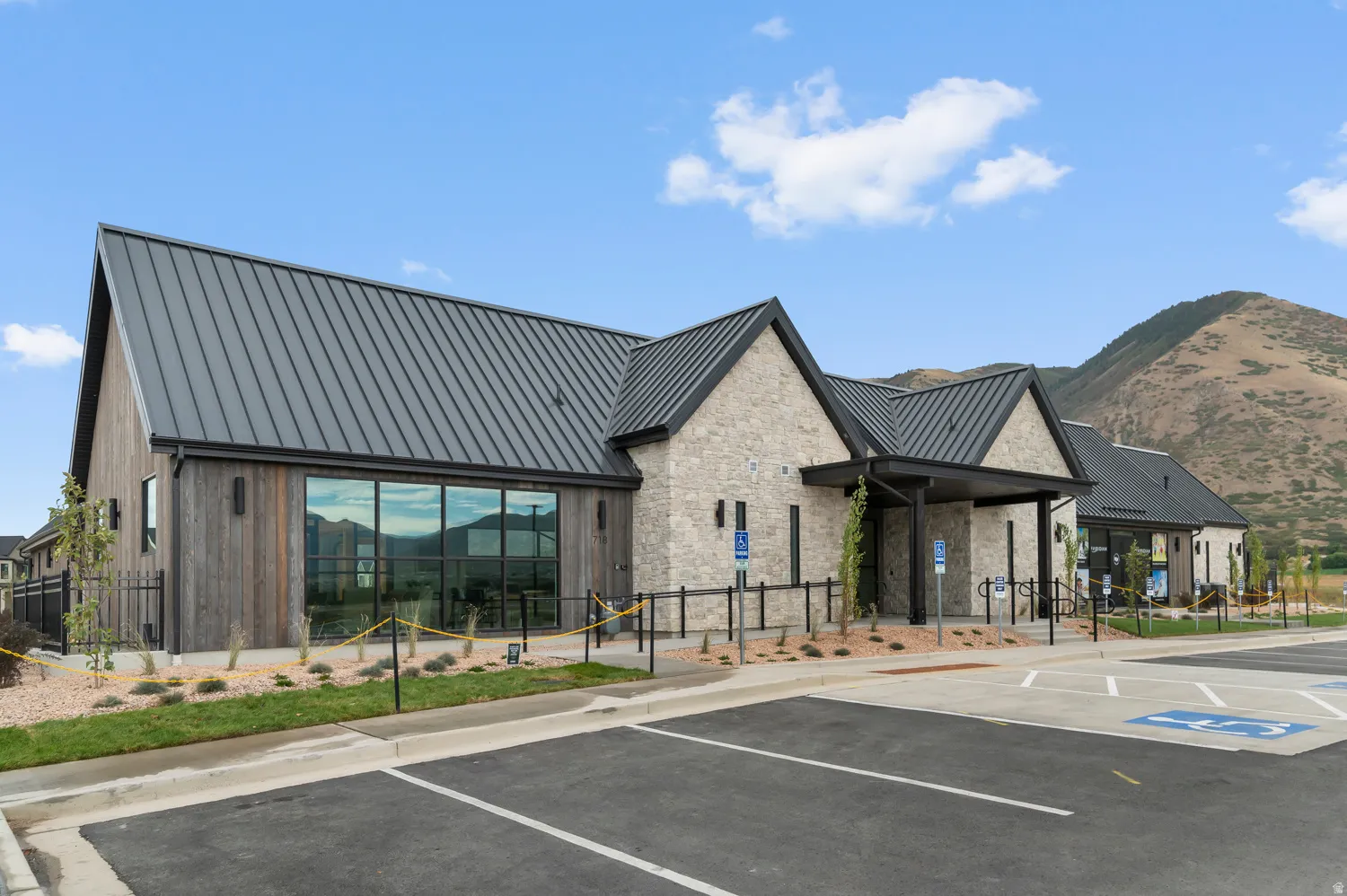 Modern architectural design featuring a prominent metal roof, stone and wood-paneled exterior, expansive storefront windows, and a paved parking area