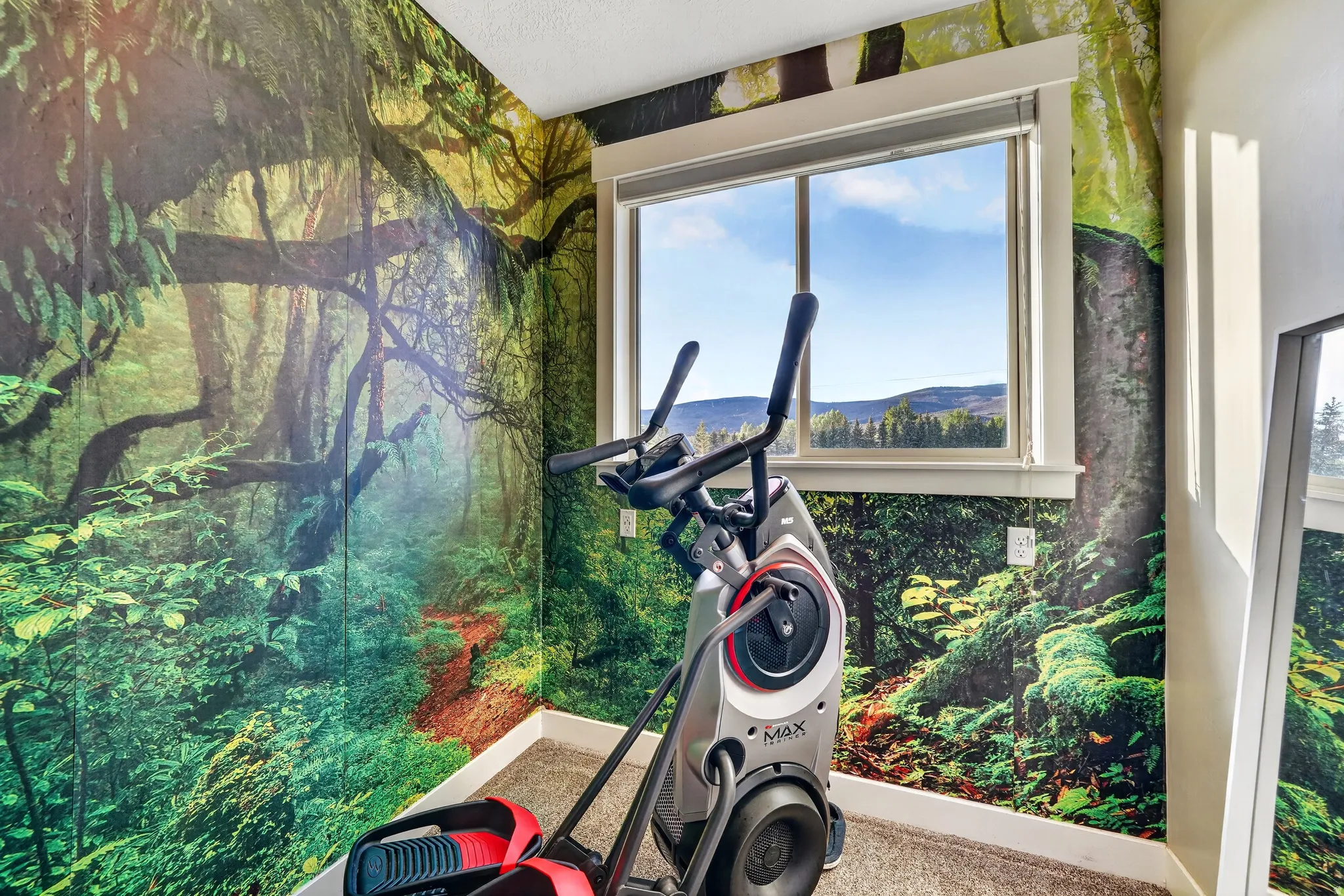 Room featuring vibrant forest-themed wall coverings, a large window, white window trim, and a mountain and sky backdrop