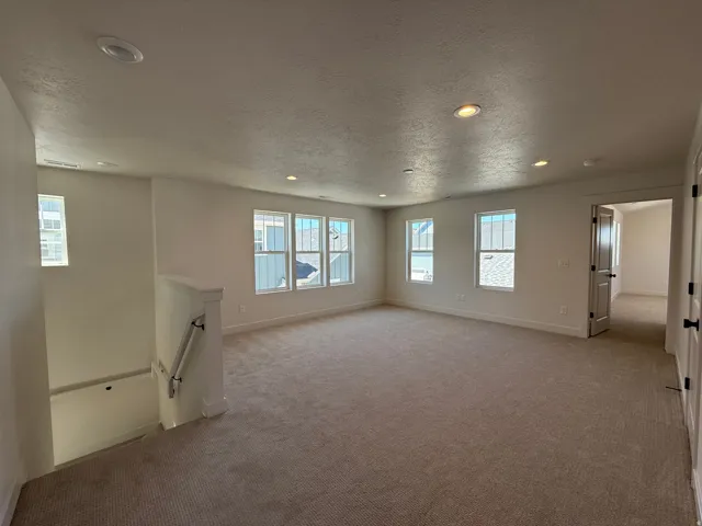 Spacious carpeted room featuring multiple windows, recessed lighting, a neutral color palette, and a staircase with white railing